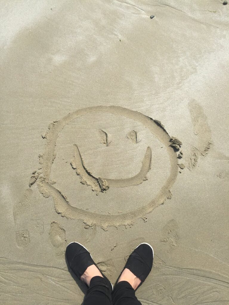 A smiley face drawn in sand on a sunny beach, captured with shoes visible.